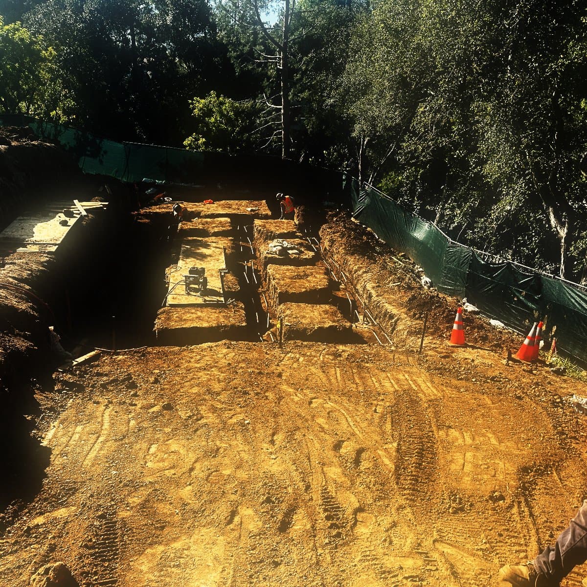 Grade beam trenches cut in a grid across an excavated hillside platform, the rock cut face standing along one side and screened perimeter fencing along the other