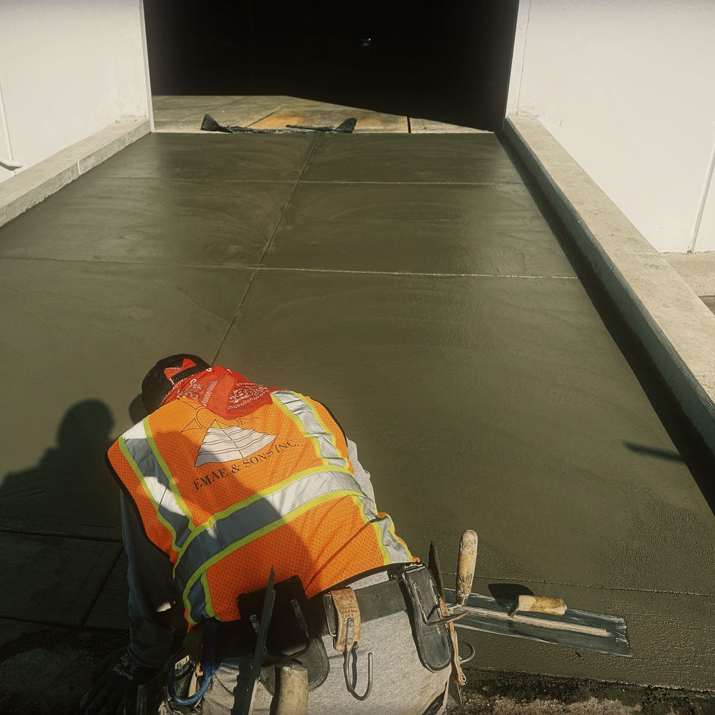 A concrete finisher in an EMAE & Sons Inc. hi-vis vest kneeling on the freshly placed apron at Costa Mesa, hand troweling the surface, with a fresno and edging tools resting on the slab beside him