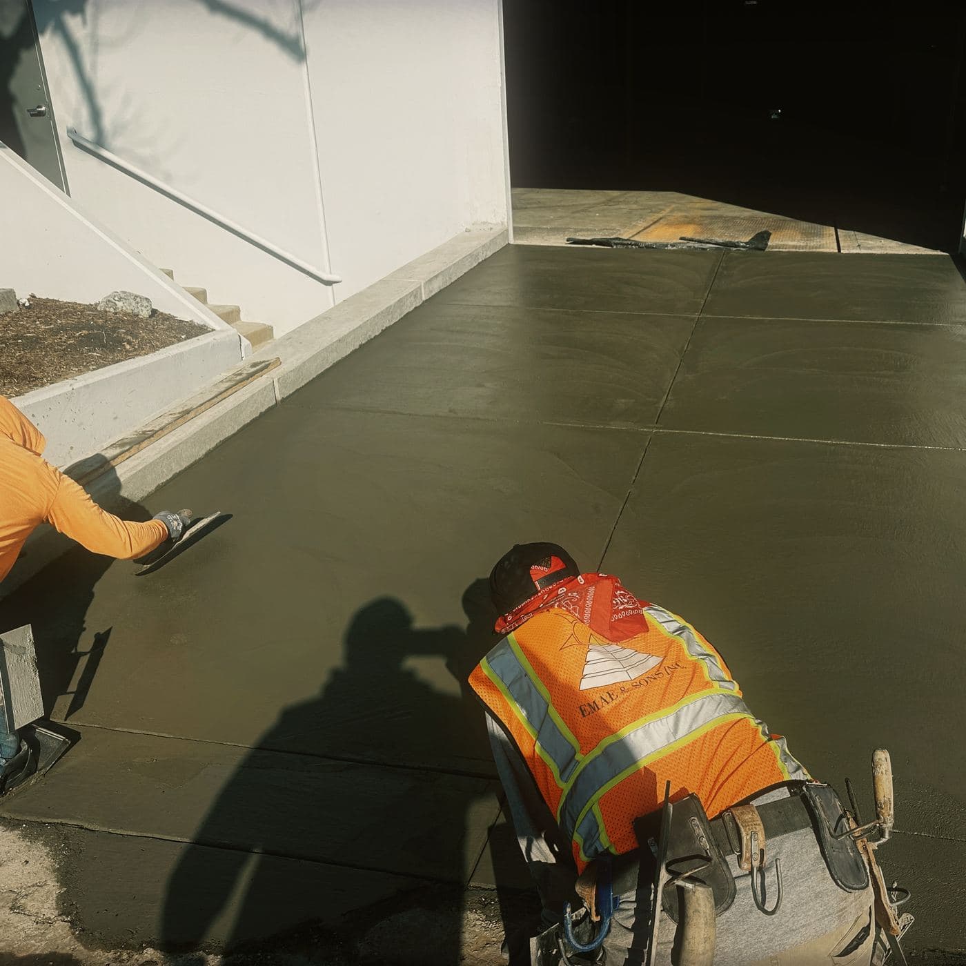 Two EMAE & Sons finishers working the same wet concrete apron at Costa Mesa, one troweling at the center and one floating the edge against the formed curb
