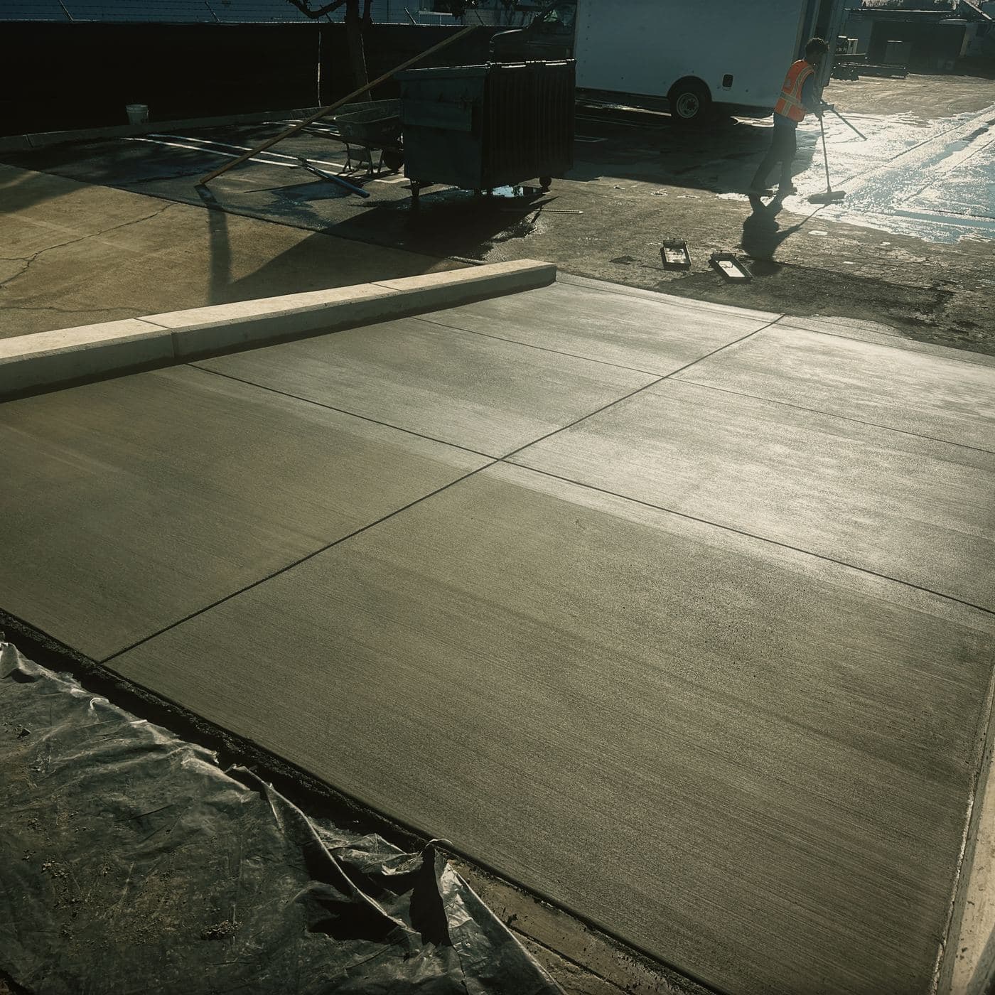 The second, wider concrete apron at Costa Mesa with a broom finish and a tooled joint grid, bounded by a poured curb, with a crew member squeegeeing wash water off the lot behind it