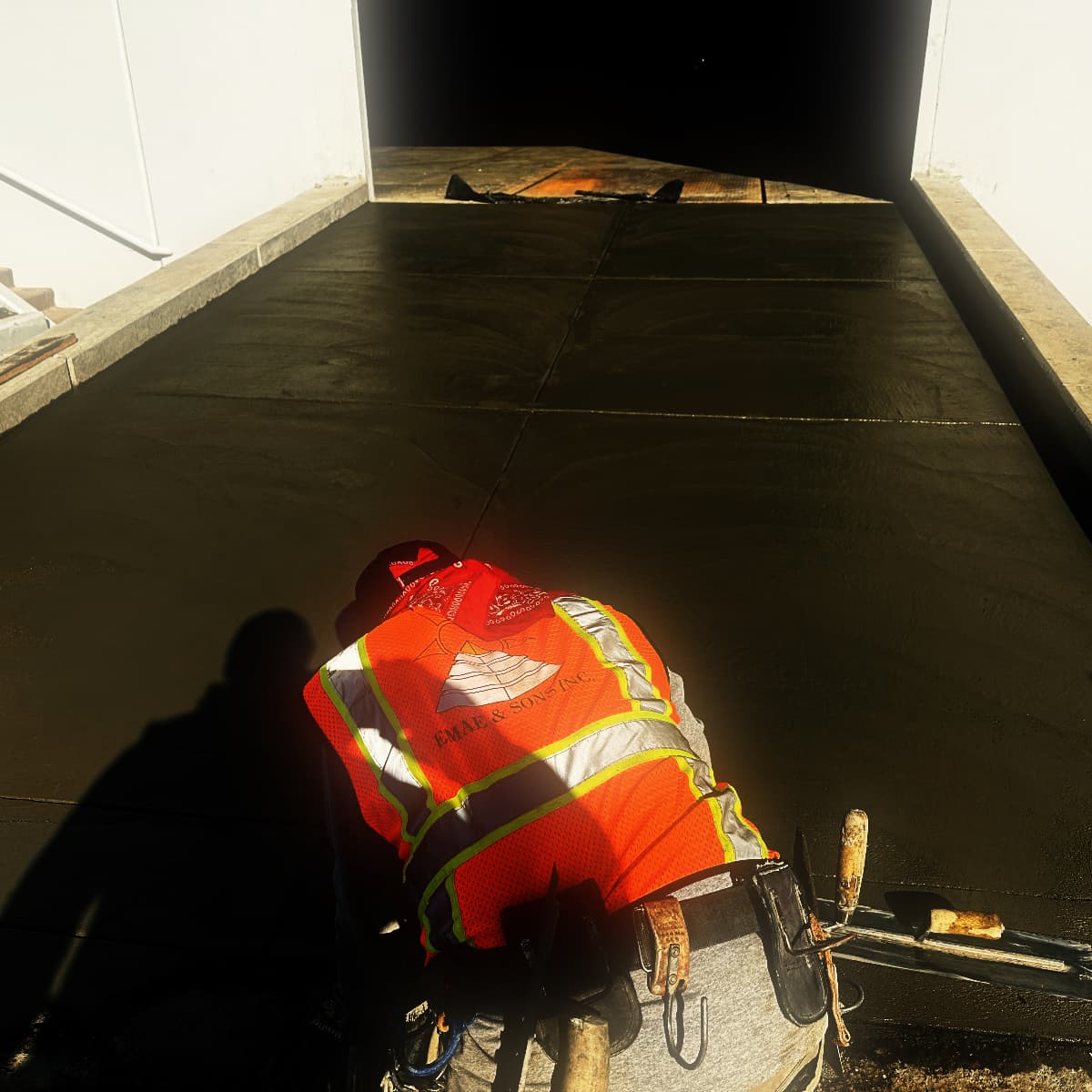 A finisher in a high-visibility vest kneeling to steel-trowel the edge of a freshly placed concrete apron, hand tools laid on the slab beside him
