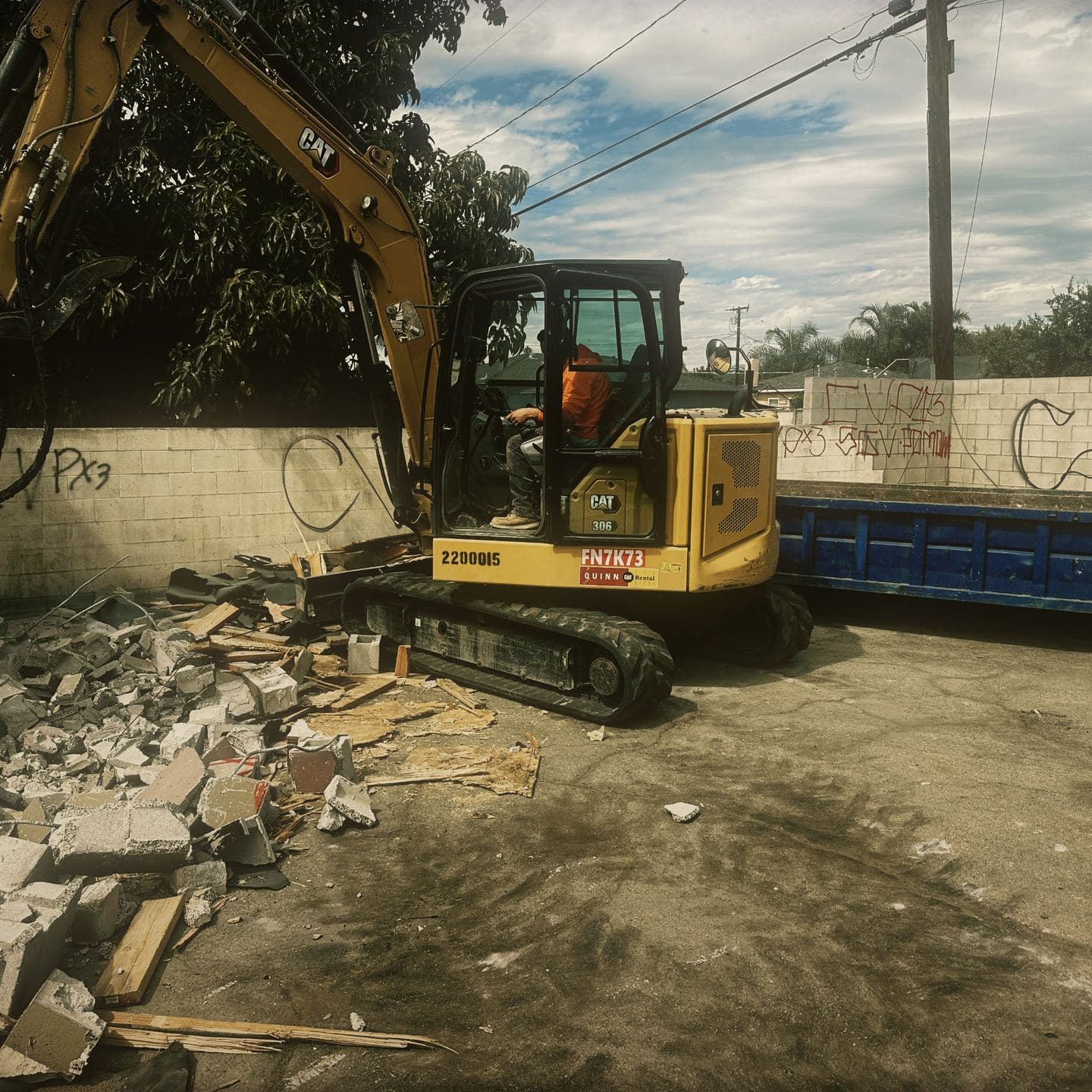A tracked excavator fitted with a hydraulic breaker standing over broken masonry in Pomona, the debris container alongside it and the tagged boundary wall behind