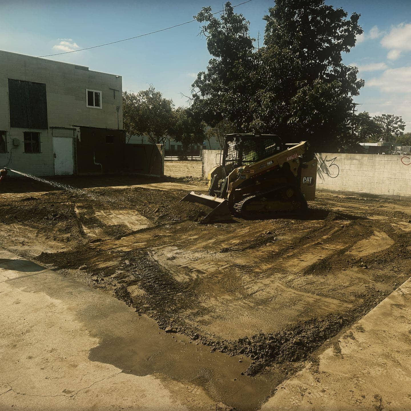 Water arcing from a hose across the freshly graded lot in Pomona for dust control, with a compact track loader working the ground beyond