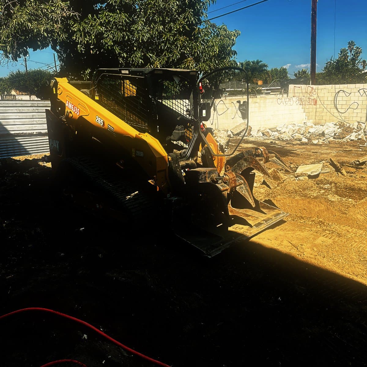 A compact track loader fitted with a grapple bucket standing on a cleared commercial lot, broken concrete stacked along the boundary wall behind it