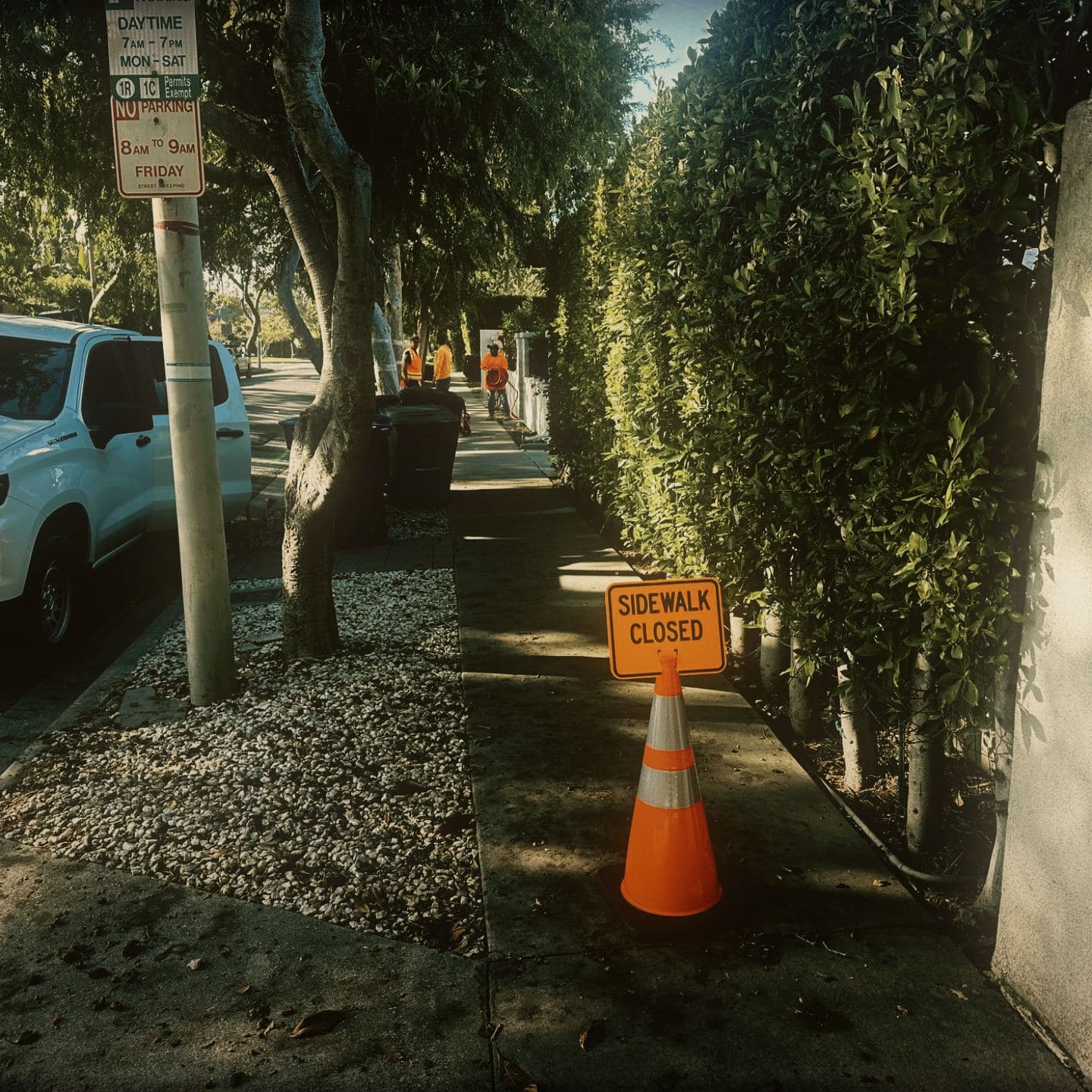 A SIDEWALK CLOSED sign mounted on a cone at the approach to the intact sidewalk, with crew working further down the block