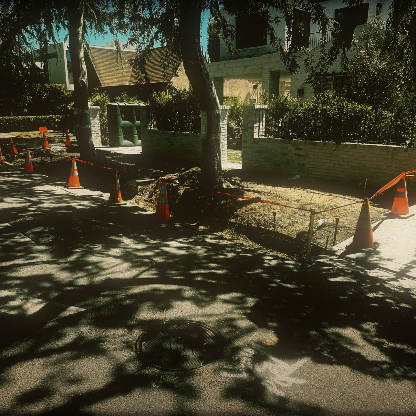 The right-of-way strip excavated back to the property wall, held behind a line of cones and red barrier tape, with grade stakes set in the open ground