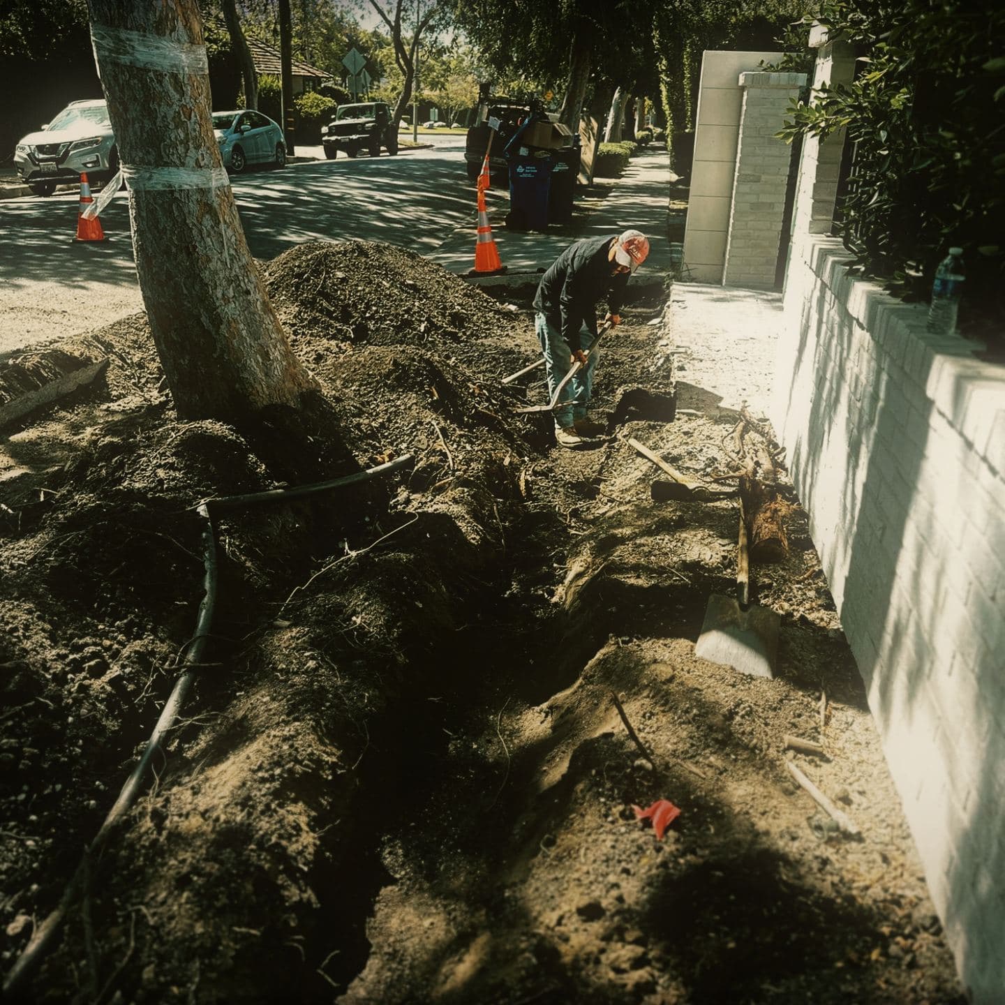 A crew member dressing the trench line by hand with a shovel alongside the property wall, black conduit already laid in the bottom of the trench