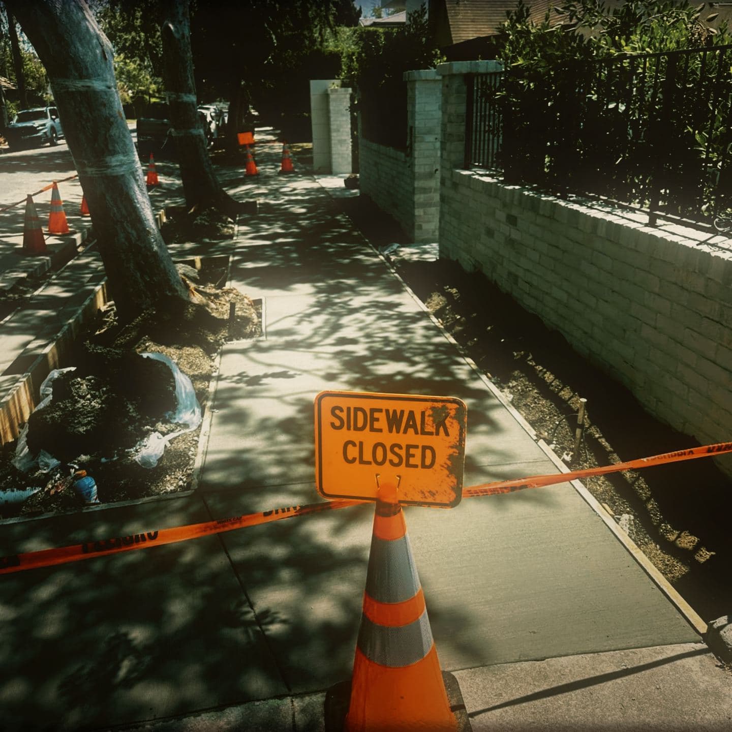 A SIDEWALK CLOSED sign and danger tape held across the newly poured sidewalk at Westmount, the finished run receding down the block behind a cone taper