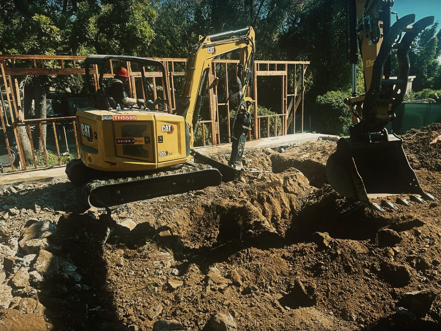 An EMAE & Sons operator working the company's Caterpillar 305 mini excavator, trenching beside timber formwork on a Brentwood hillside