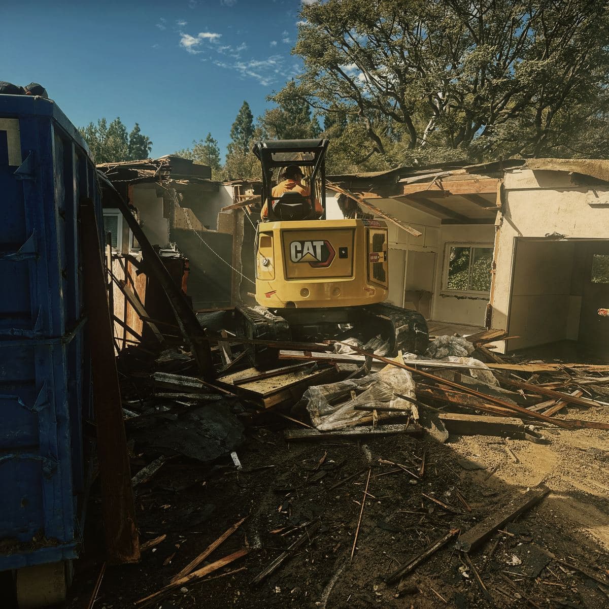 A tracked excavator taking down the wall of a single-story stucco house on the Brentwood lot, with a roll-off bin alongside and broken timber across the ground