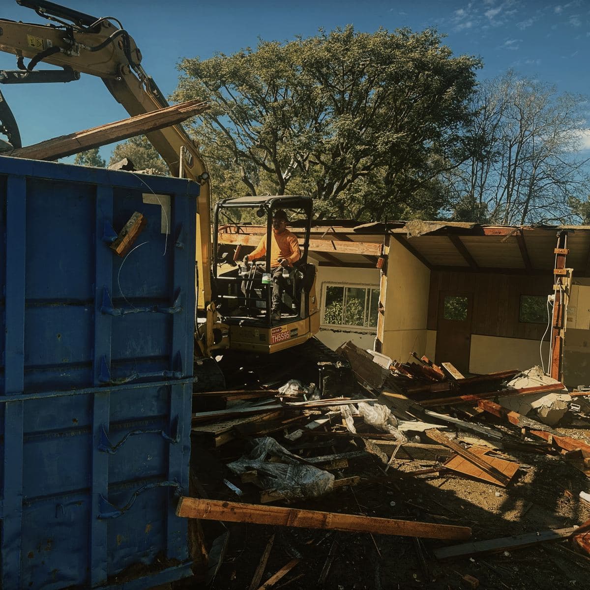 An excavator loading demolition timber directly into a roll-off bin on the Brentwood site, the remaining half of the house still standing behind it