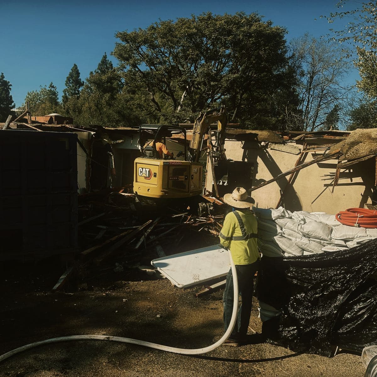 A crew member in a hi-vis vest running a hose across the demolition area on the Brentwood site to suppress dust, with a roll-off bin and the part-demolished house behind