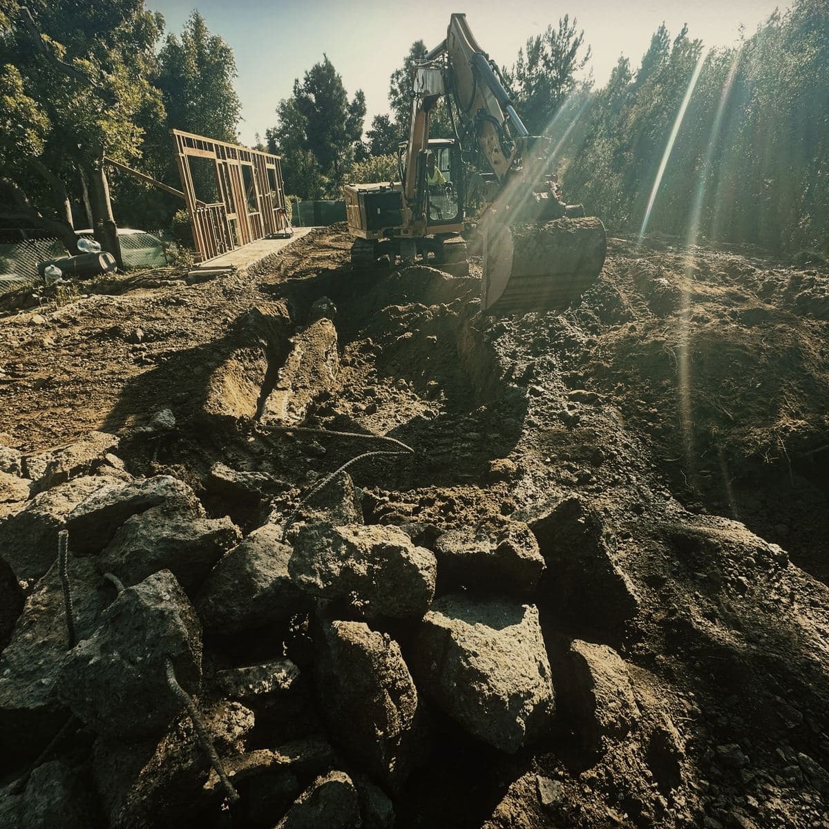 Broken rock stacked across the excavated Brentwood hillside platform in pieces up to boulder size, with an excavator working the face beyond