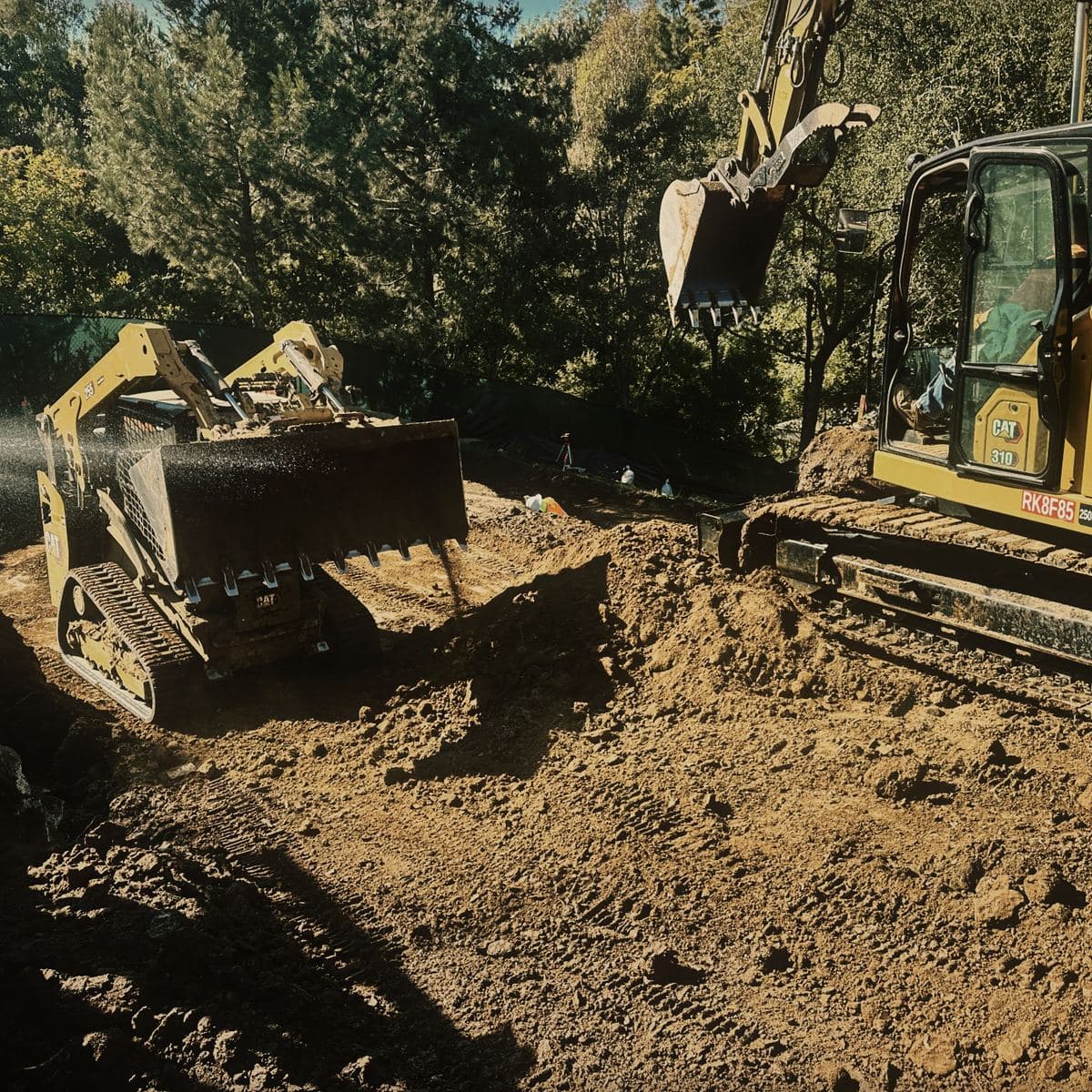 A compact track loader working the excavated surface on the Brentwood site under a spray of water, with a tracked excavator alongside it