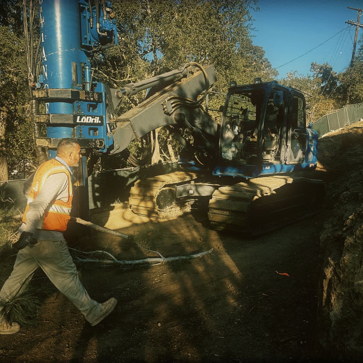 A tracked mast drilling rig set up on the Brentwood hillside platform to drill caissons, with the cut face and screened perimeter fence behind it