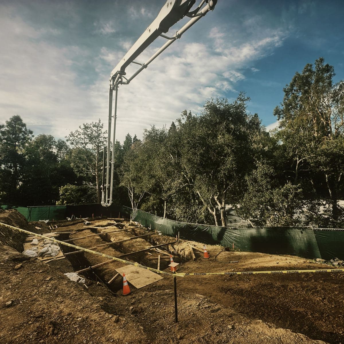 A concrete boom pump reaching over the excavated Brentwood platform to place the grade beams, the formed runs taped off below it