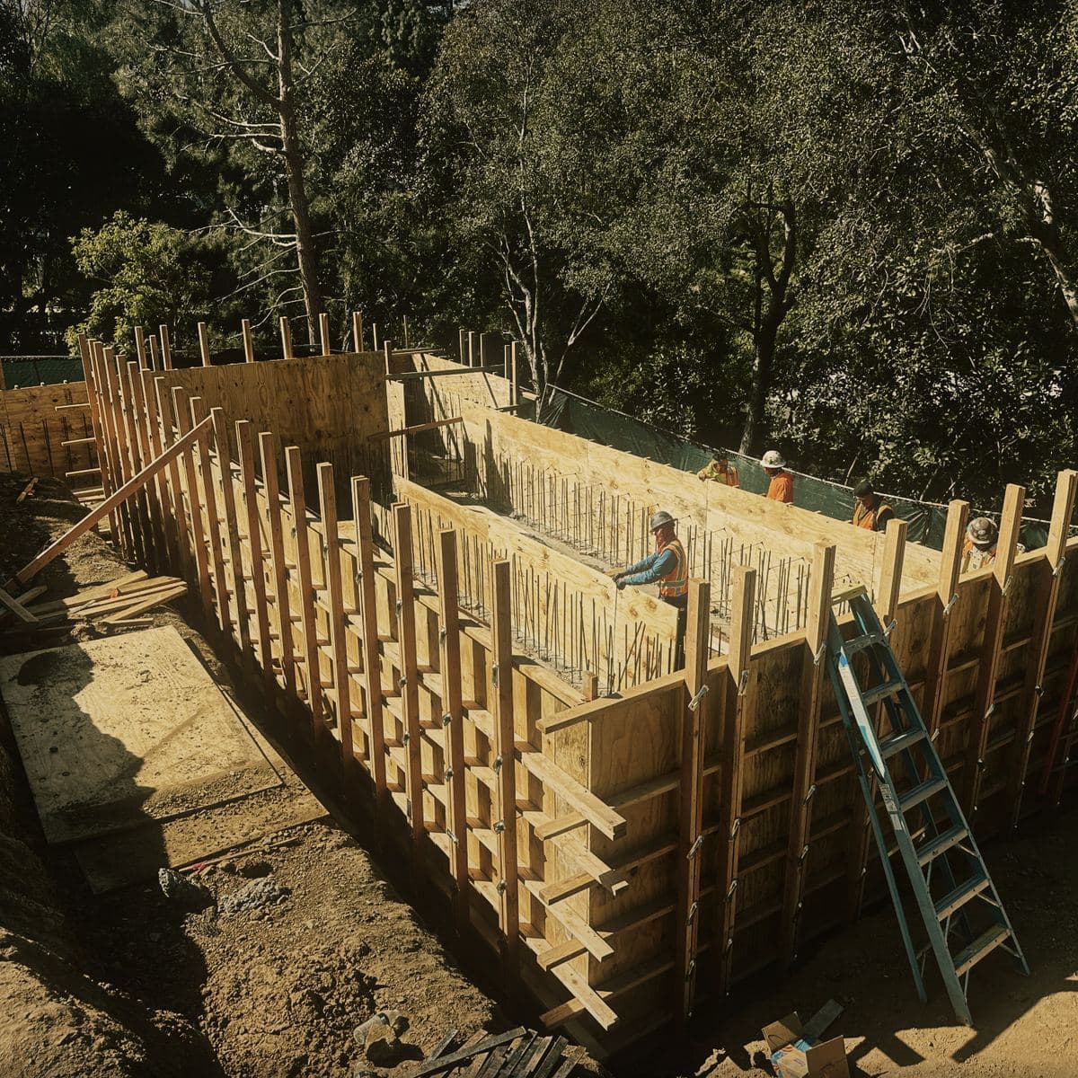 Timber wall forms standing the full depth of the Brentwood basement excavation, braced and laddered, running the length of the cut face