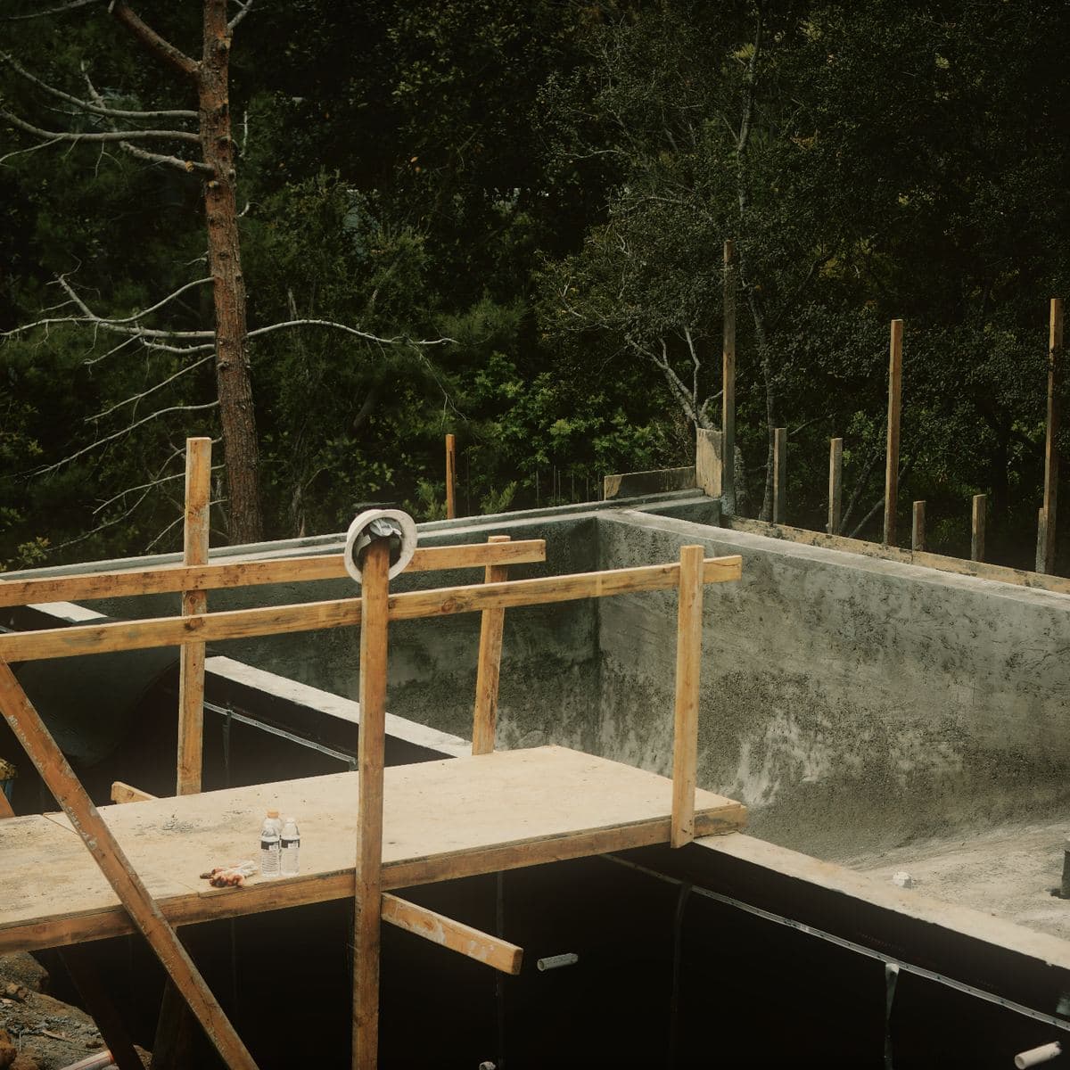 The stripped concrete basement and pool structure on the Brentwood site with its external face covered in black waterproofing membrane and drainage board, wall dowels standing above the pour line