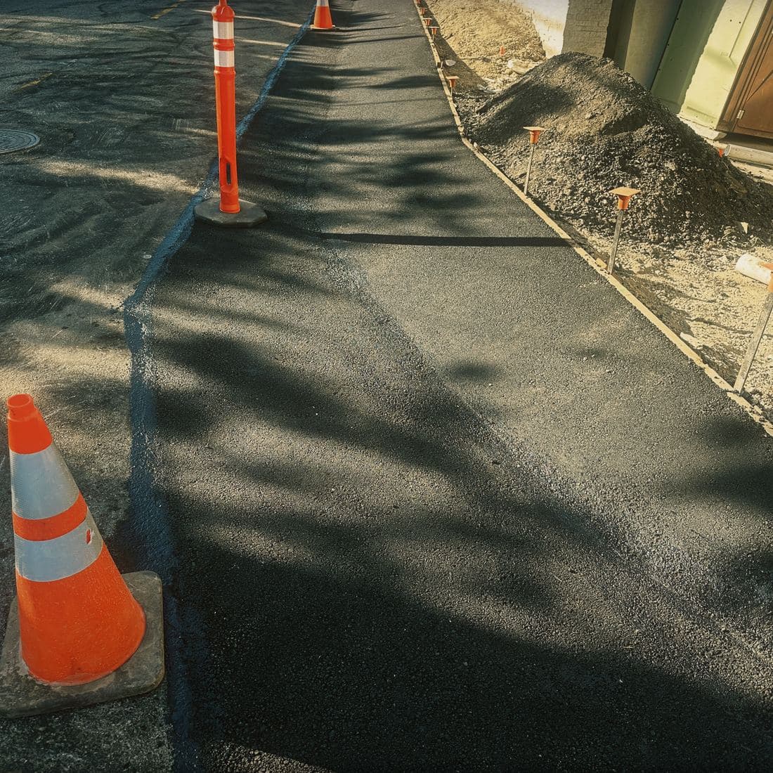 The closed lane on the Brentwood street: a traffic cone and delineator post tapering up the length of the new asphalt, with the formed edge on the far side and every steel stake capped in orange
