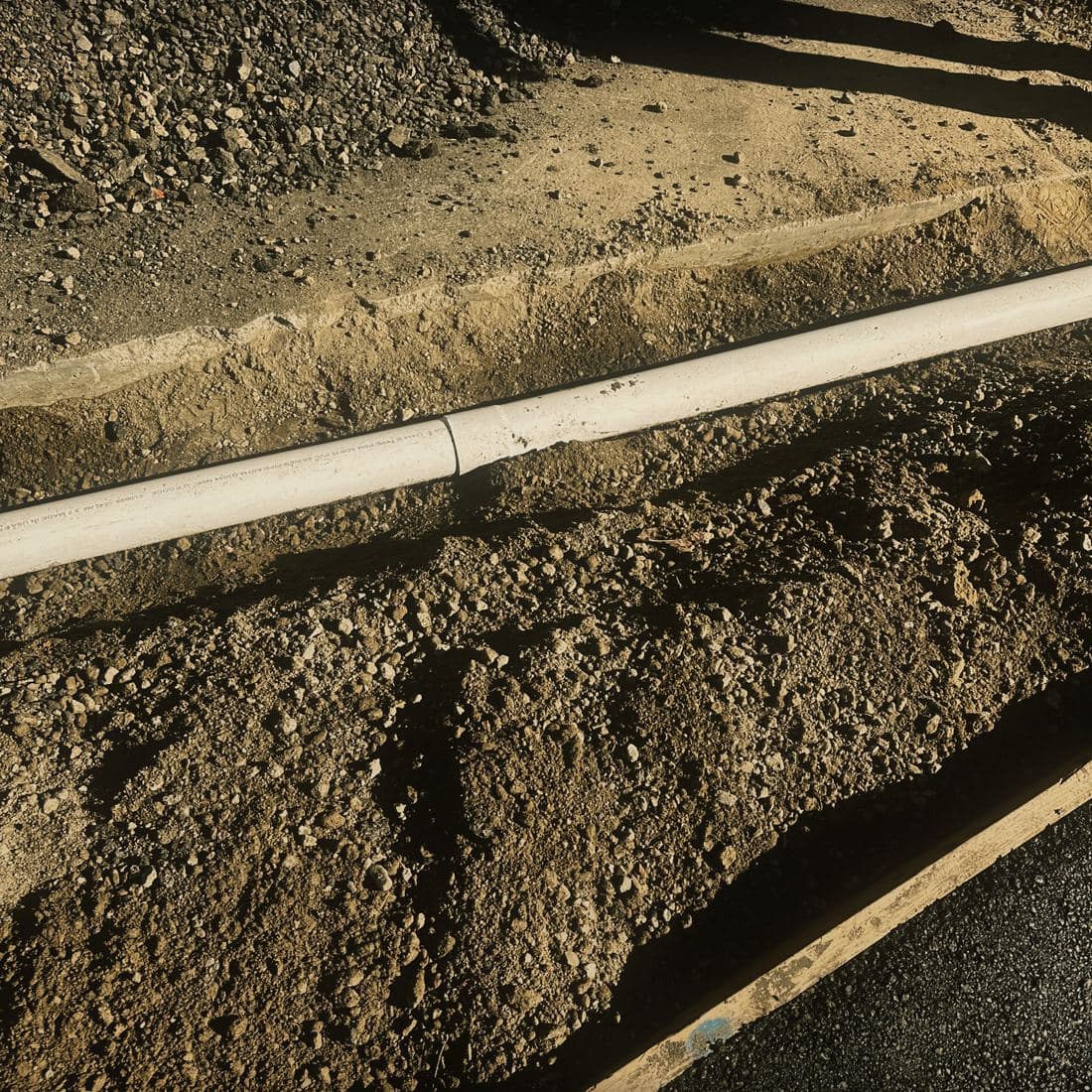 A gravity line in white SDR 35 PVC laid along the bottom of an open trench on the Brentwood street, running against the existing concrete gutter with spoil banked on the far side and a form board beyond it