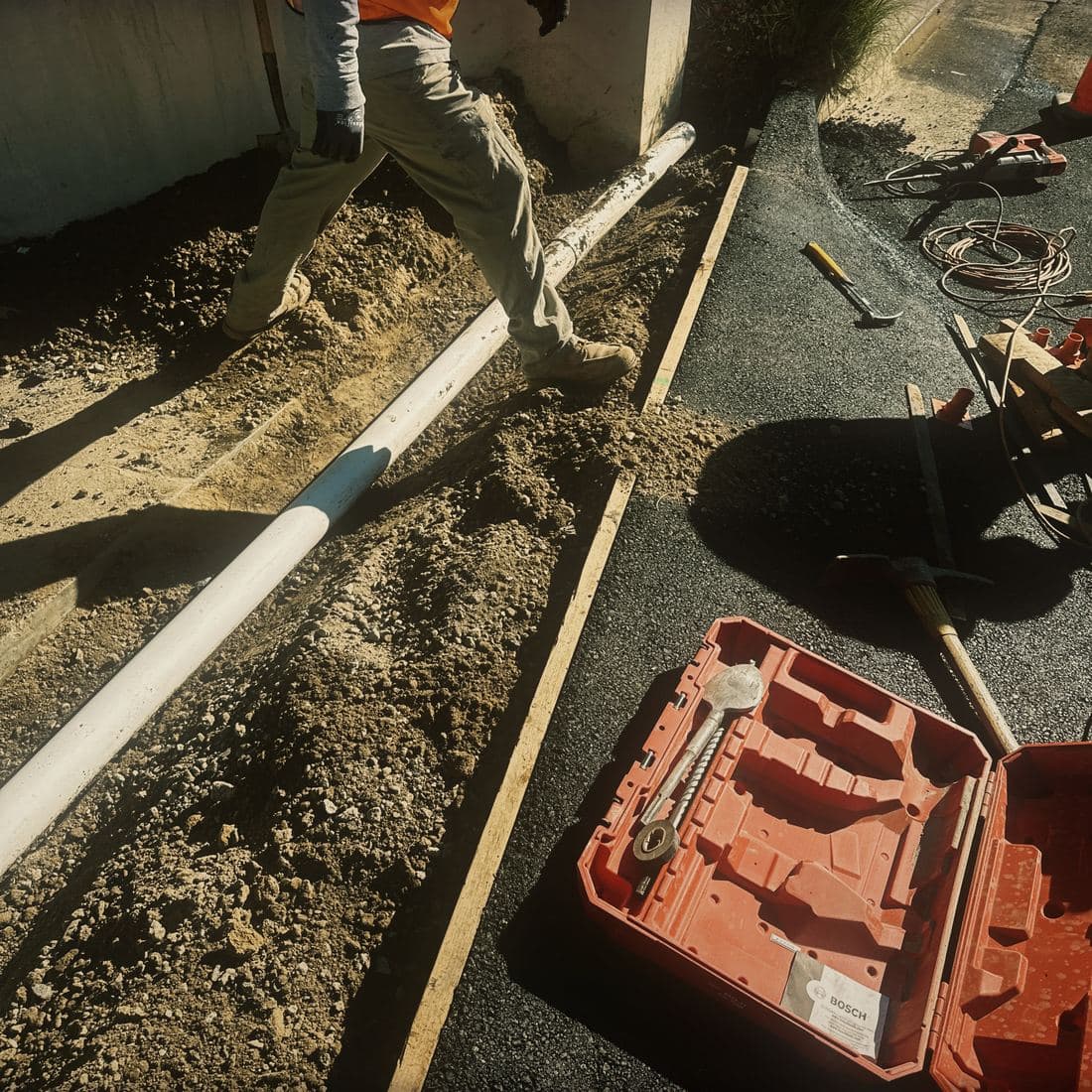 A crew member in a hi-vis vest walking the length of the open trench on the Brentwood street with the pipe run set in it, the formed edge on one side and hand tools and a breaker staged out on the new asphalt