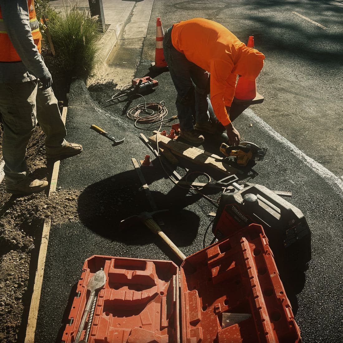 Two crew members working in the closed lane of the Brentwood street, one cutting a form board with a circular saw and one standing at the trench edge, with a traffic cone and the new asphalt around them