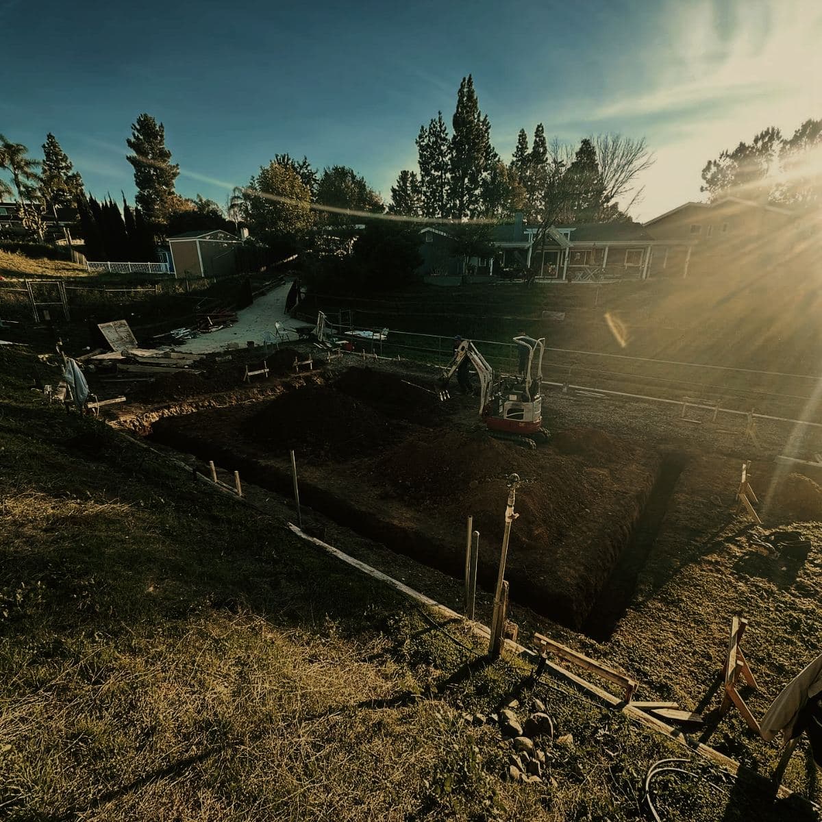 Footing trenches excavated in a back yard at sunrise, spoil heaped in the middle of the pad and a mini excavator standing at the far corner