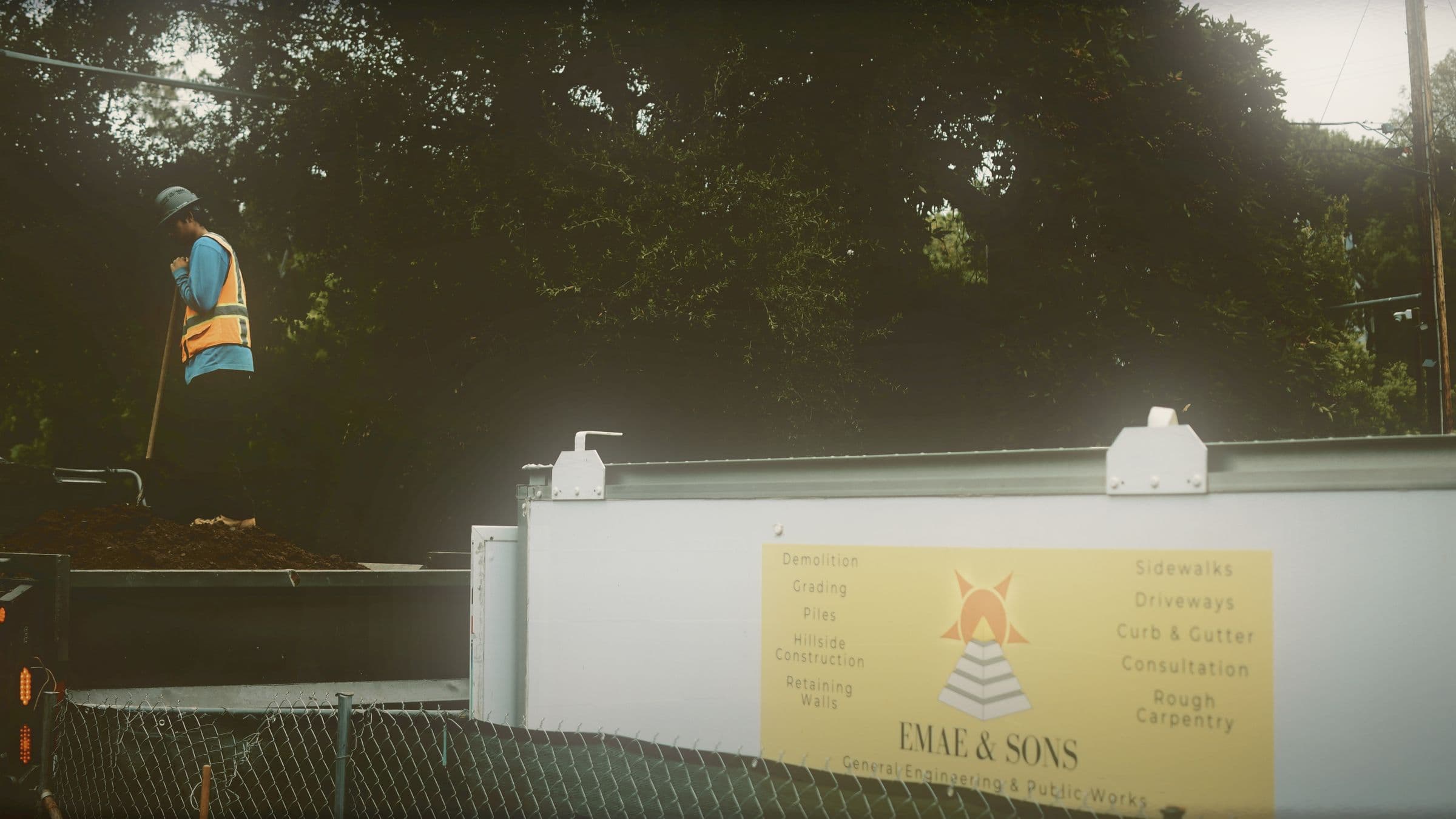 An EMAE & Sons site board on a fenced job site listing the firm's own services — demolition, grading, piles, hillside construction, retaining walls, sidewalks, driveways, curb and gutter — with a crew member working behind it